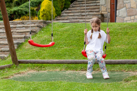One single sad lonely elementary school age girl, unhappy child sitting on a swing alone, playground, outdoors shot. No friends, sadness, loneliness concept, social issues, one person. Outdoors shotの写真素材