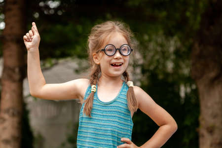 Young happy curious elementary school age girl, clever intelligent genius child student in big funny glasses has a brilliant idea, finger pointing up, eureka pose concept one person, outdoors portraitの写真素材