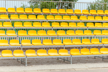 Empty vacant stadium stands, chairs, plastic yellow arena spectator seats on a small football soccer field, daytime, pan, nobody. No people, sports event site, fans absence, rows of seats outdoorsの写真素材