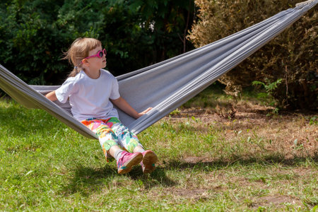 One single school age child, girl wearing cool sunglasses sitting on a hammock, resting in the sun summer leisure activities outdoors lifestyle scene. One person alone, copy space, nature, backyardの写真素材