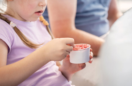 Anonymous elementary school age child girl eating ice cream from a small white blank paper cup with a spoon, closeup, detail, one person. Kids eating sweet food, nutrition, sugar simple conceptの写真素材