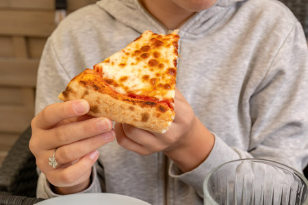 Child holding a slice of margherita cheese pizza close up eating meal, mozzarella topping, casual dining, hands with pizza slice cozy atmosphere, face obscured, warm baked food, meal enjoyment conceptの写真素材