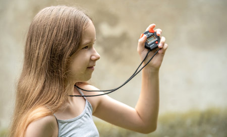 Young girl holding a stopwatch measuring time outdoors child timing event, sports training, concentration, time management, stopwatch in hand, childhood learning, outdoor activity sport timing conceptの写真素材