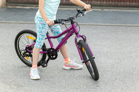 Young anonymous child cyclist girl kid biker sitting on a purple bicycle standing on the street, asphalt road, hands on handlebars, foot on pedal, learning to ride outdoor recreation scene, copy spaceの写真素材