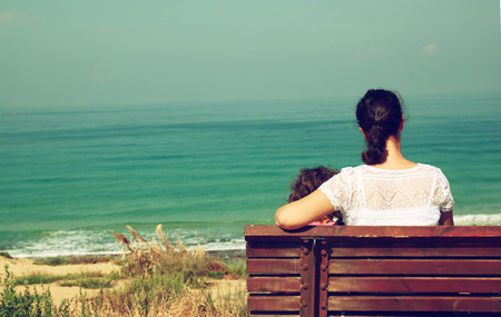 mother and child sitting on bench and looking forward at the seaの写真素材