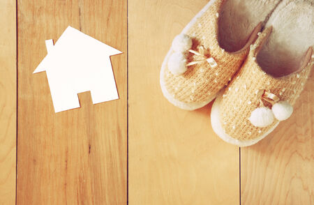 top view of warm woman slippers over wooden floor and paper house shape as welcome home conceptの写真素材