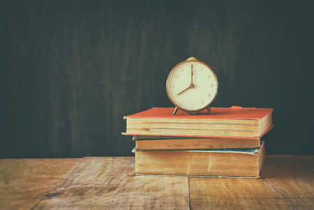 old clock over stack of books and wooden table background. retro faded style imageの写真素材