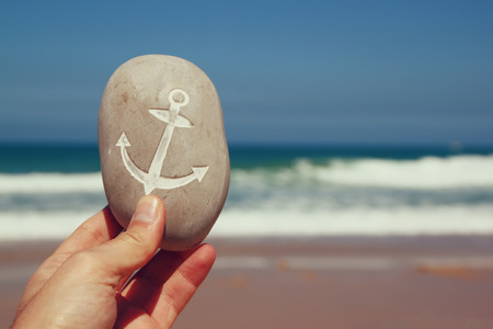 man hand holding one stone pebbles with the anchor sign  against sandy beach and sea horizonの写真素材
