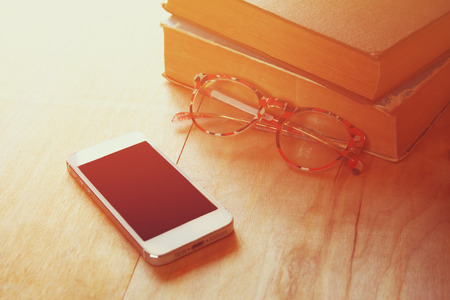 reading glasses, stack of old books and smart phone over wooden table, retro filtered imageの写真素材