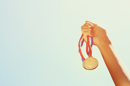 woman hand raised, holding gold medal against sky