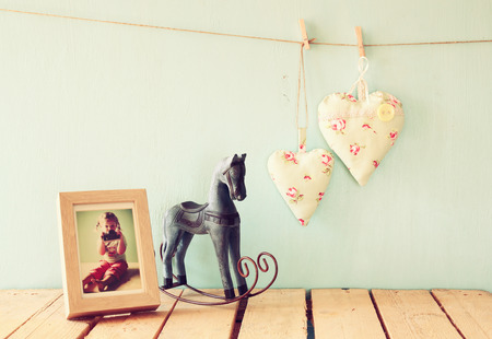 vintage rocking horse next to fabric hearts hanging on the rope on wooden floor and photo frame with kid's old photography. retro filtered imageの写真素材