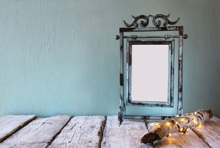 low key image of old victorian steel blue blank frame next to log tree with fairy christmas lights on wooden table. faded retro filtered imageの写真素材