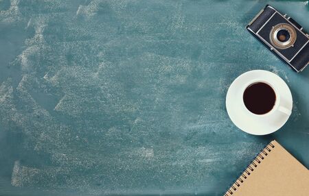 top view image of  cup of coffee, notebook and vintage camera over blackboard background