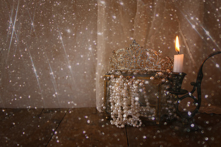 low key image of beautiful diamond queen crown, white pearls next to burning candle. vintage...