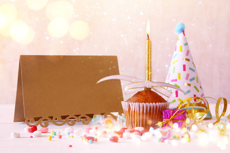 Birthday concept with cupcake and candle next to empty greeting card on wooden table. Glitter lights overlayの写真素材