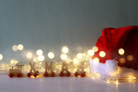 Low key Image of warm garland lights next to santa hat and wooden letters spelling word: XMAS