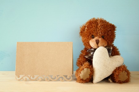 Cute teddy bear sitting and holding a heart next to blank card, on wooden tableの写真素材