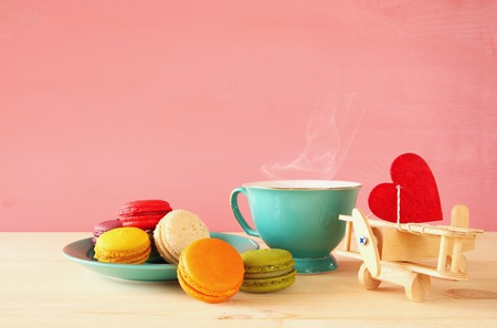 Mint vintage cup of coffee and colorful macaron or macaroon next to toy plane with heart on wooden table over pink backgroundの写真素材