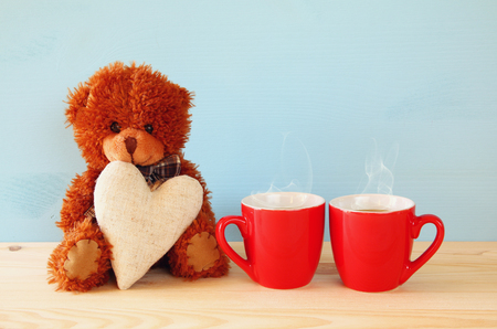 Valentines day concept. Cute teddy bear sitting and holding a heart next to couple of coffee cups, on wooden tableの写真素材