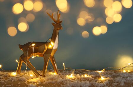 Gold shiny reindeer on snowy wooden table with christmas garland lightsの写真素材