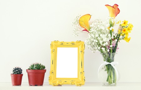 Image of beautiful bouquet of yellow spring flowers next to blank vintage photo frame over white table. For photography mock up montageの写真素材