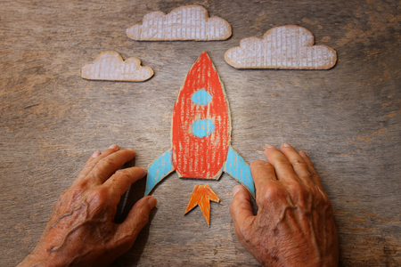 A close up of an elderly man holding a paper rocket on a wooden table. Concept of thinking about childhood dreams, sadness and loneliness.の写真素材