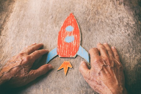 A close up of an elderly man holding a paper rocket on a wooden table. Concept of thinking about...