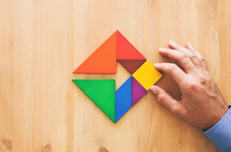 man's hand holding a missing piece in a square tangram puzzle, over wooden tableの写真素材