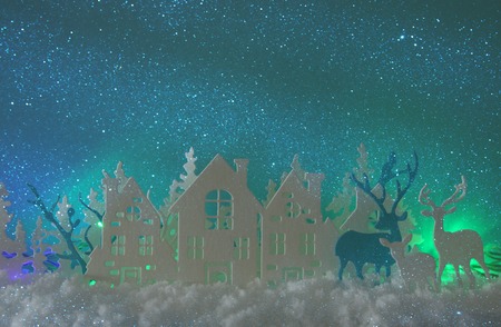 Magical Christmas paper cut winter background landscape with houses, trees, deer and snow in front of northern lights backgroundの写真素材