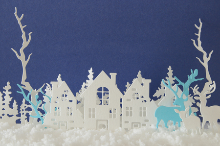 Magical Christmas paper cut winter background landscape with houses, trees, deer and snow in front of blue backgroundの写真素材