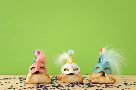 Purim celebration concept (jewish carnival holiday). Traditional hamantaschen cookies with cute clown hats and masks over wooden table and green backgroundの写真素材