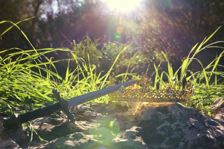 mysterious and magical photo of silver king crown and sword over the stone in the England woods or field landscape with light flare. Medieval period conceptの写真素材