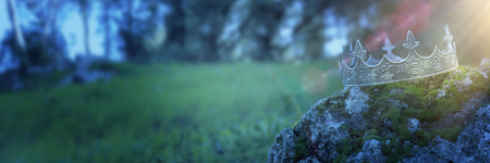 Mysterious and magical photo of silver king crown over the stone covered with moss in the England woods or field landscape with light flare. Medieval period conceptの写真素材