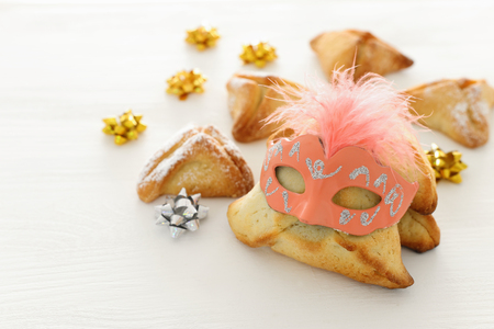 Purim celebration concept (Jewish carnival holiday). Traditional hamantaschen cookies with cute mask over white wooden tableの写真素材