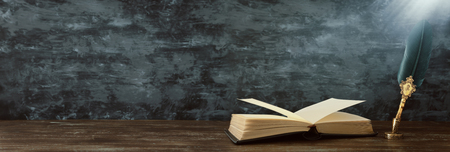 Old feather quill ink pen with inkwell and old books over wooden desk in-front of black wall background. Vintage old style and filtered. Conceptual photo on history, fantasy, education and literature topicの写真素材
