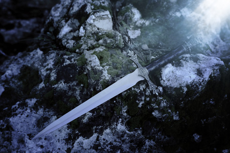 mysterious and magical photo of silver sword over the stone covered with moss in the England woods. Medieval period concept/の写真素材