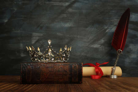 low key image of beautiful queen/king crown on the old books and feather quill ink pen over wooden...