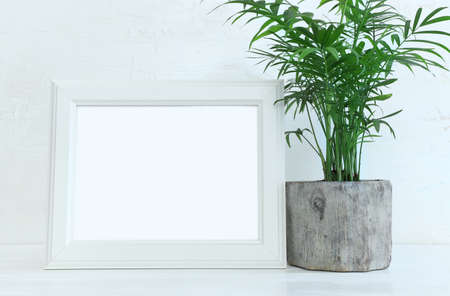 Image of white empty photo frame next to home plant over wooden table. For mockup, can be used for...