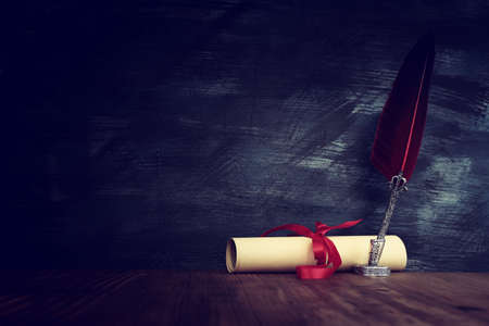 Old feather quill ink pen with inkwell and paper scroll over wooden desk in front of black wall background. Conceptual photo on history, fantasy, education and literature topic.の写真素材