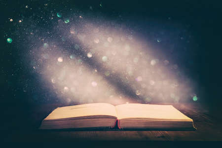 Magical image of open antique book over wooden table with glitter overlay