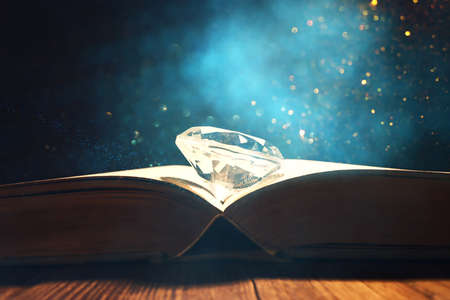 image of open antique book and diamond on wooden table with glitter overlayの写真素材