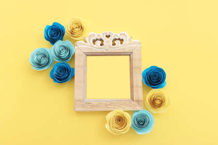 Top view image of paper flowers composition and empty wooden photo frame over yellow pastel...