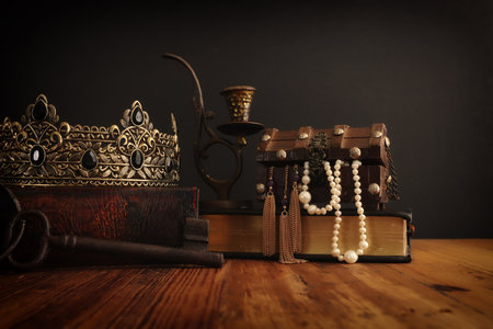 low key image of beautiful queen or king crown over antique book next to wooden treasure chest....