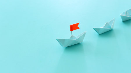 Paper boats following ahead of the leader with red flagの写真素材