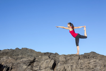 Young attractive woman doing dancers yoga pose on rocks against blue skyの写真素材