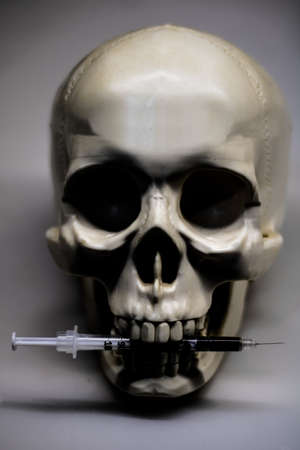 Front view of a human skull, with a cocaine syringe between its teeth, representing death from drug...