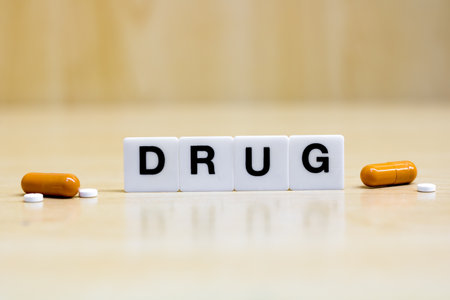 A row of small white plastic tiles, containing the letters forming the word drug, placed on a table together with tablets and capsules, to represents the concept of drug use or topic.の写真素材