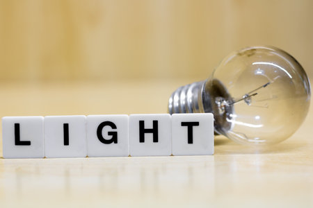 A row of small white plastic tiles, containing the letters forming the word light, placed on a table together with a light bulb, to represent the concept of artificial lighting.の写真素材