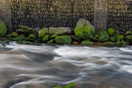 Long exposure of a flowing freshwater river with a stone wall in the backgroundの写真素材