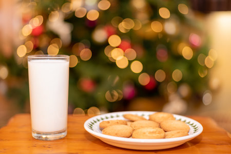 Close up of a glass of milk and a plate of cookies on a table with an illuminated christmas tree in the backgroundの写真素材